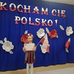 dziewczynka stoi z dyplomem na tle napisu i czerwono białych kwiatów.jpg