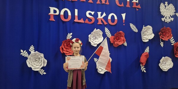 dziewczynka stoi z dyplomem na tle napisu i czerwono białych kwiatów.jpg