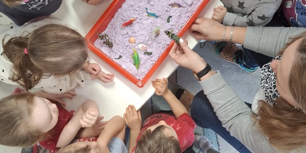 dzieci patrzą na pojemnik z kolorowym piaskiem w którym leżą dinozaury.jpg