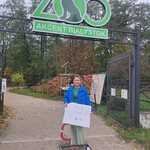 kobieta w niebieskiej kurtce stoi przed wejściem do zoo przed nią stoi skrzynka z kasztanami.jpg